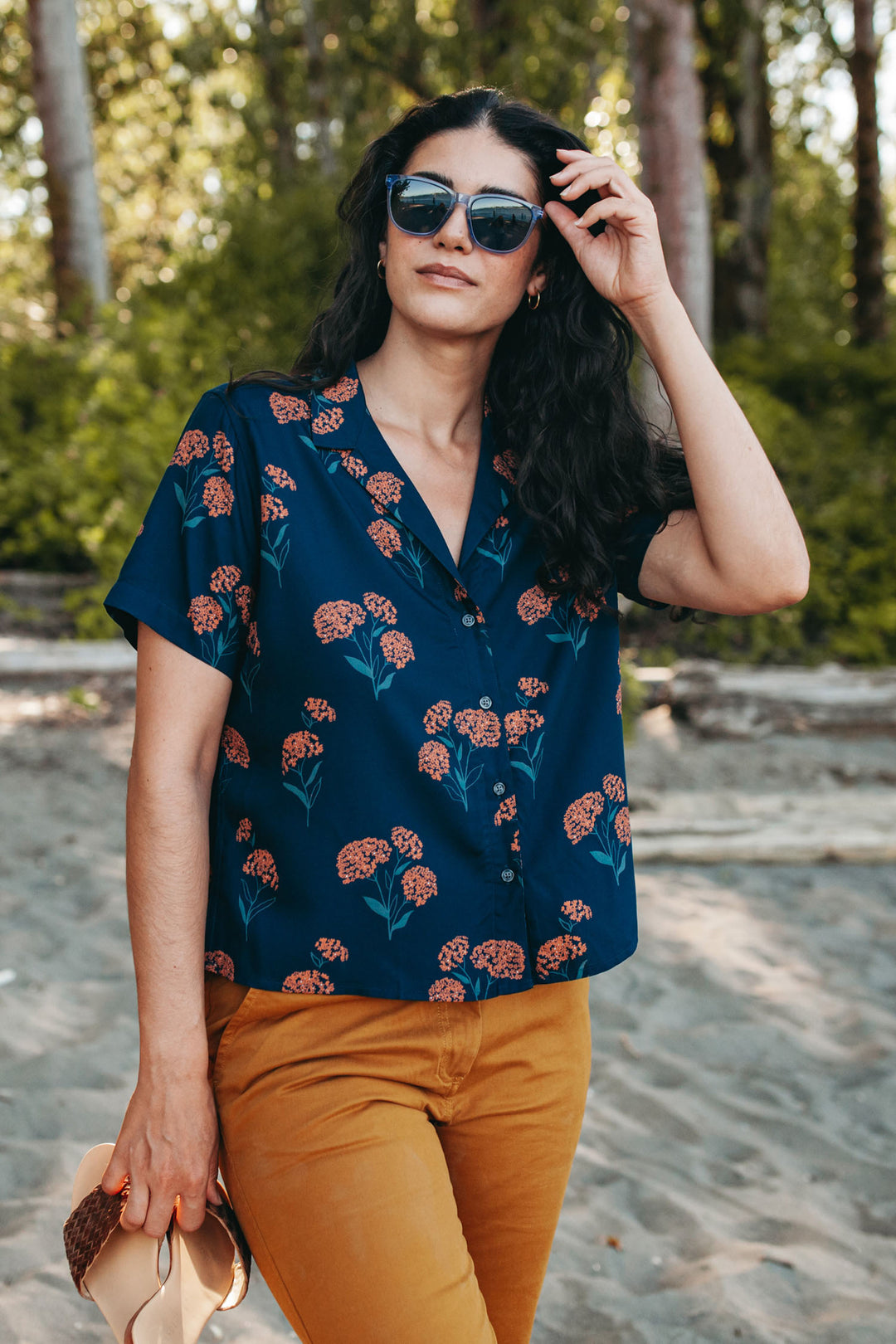 A woman in sunglasses and the Bridge & Burn Mila Cropped Shirt in Wildflower stands on a sandy path surrounded by greenery, holding a hat in her left hand and adjusting her sunglasses with her right.
