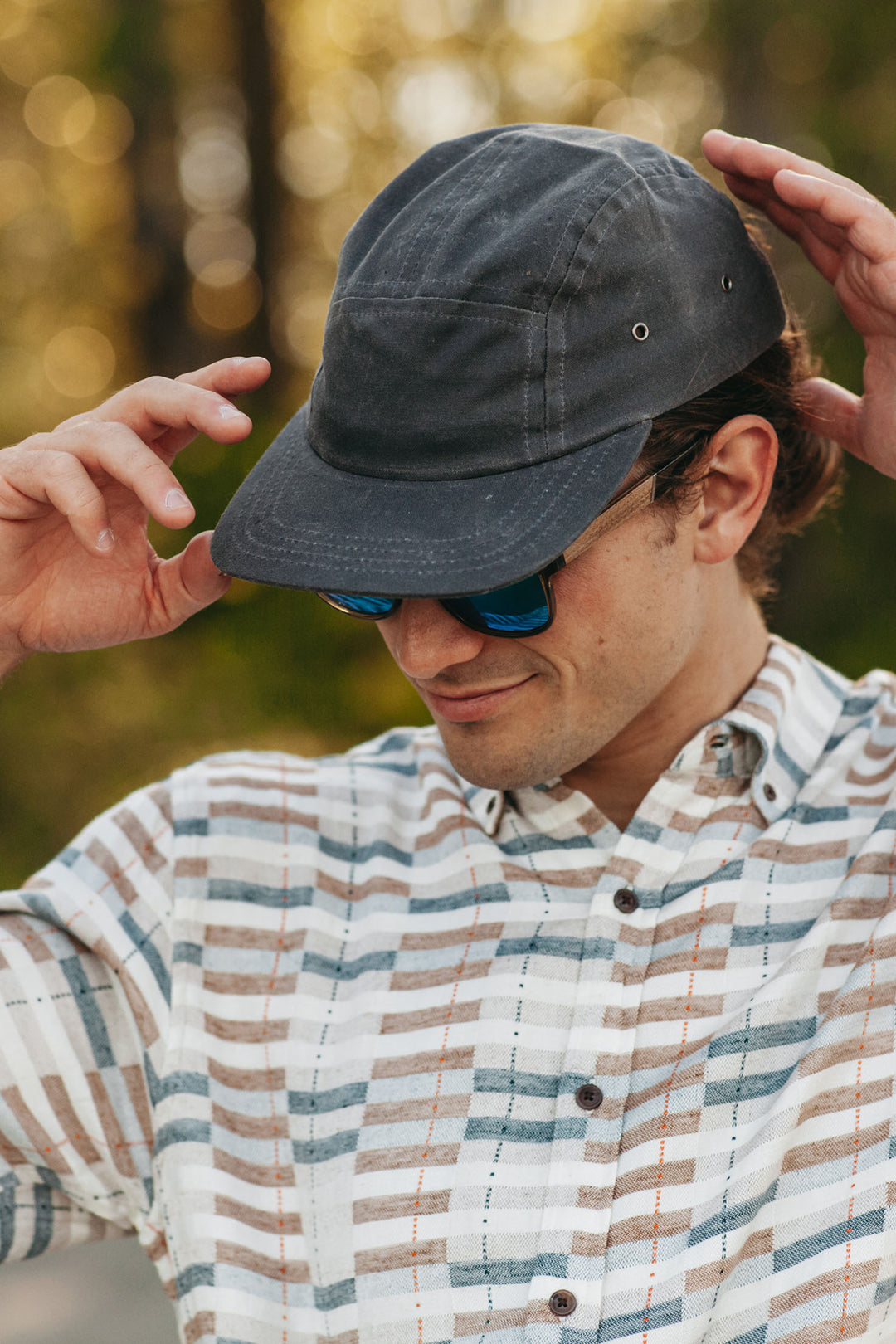 A man adjusts his Bridge & Burn Camper Waxed 5 Panel Cap in Charcoal outdoors, wearing blue sunglasses and a striped button-up shirt amid blurred trees and sunlight.