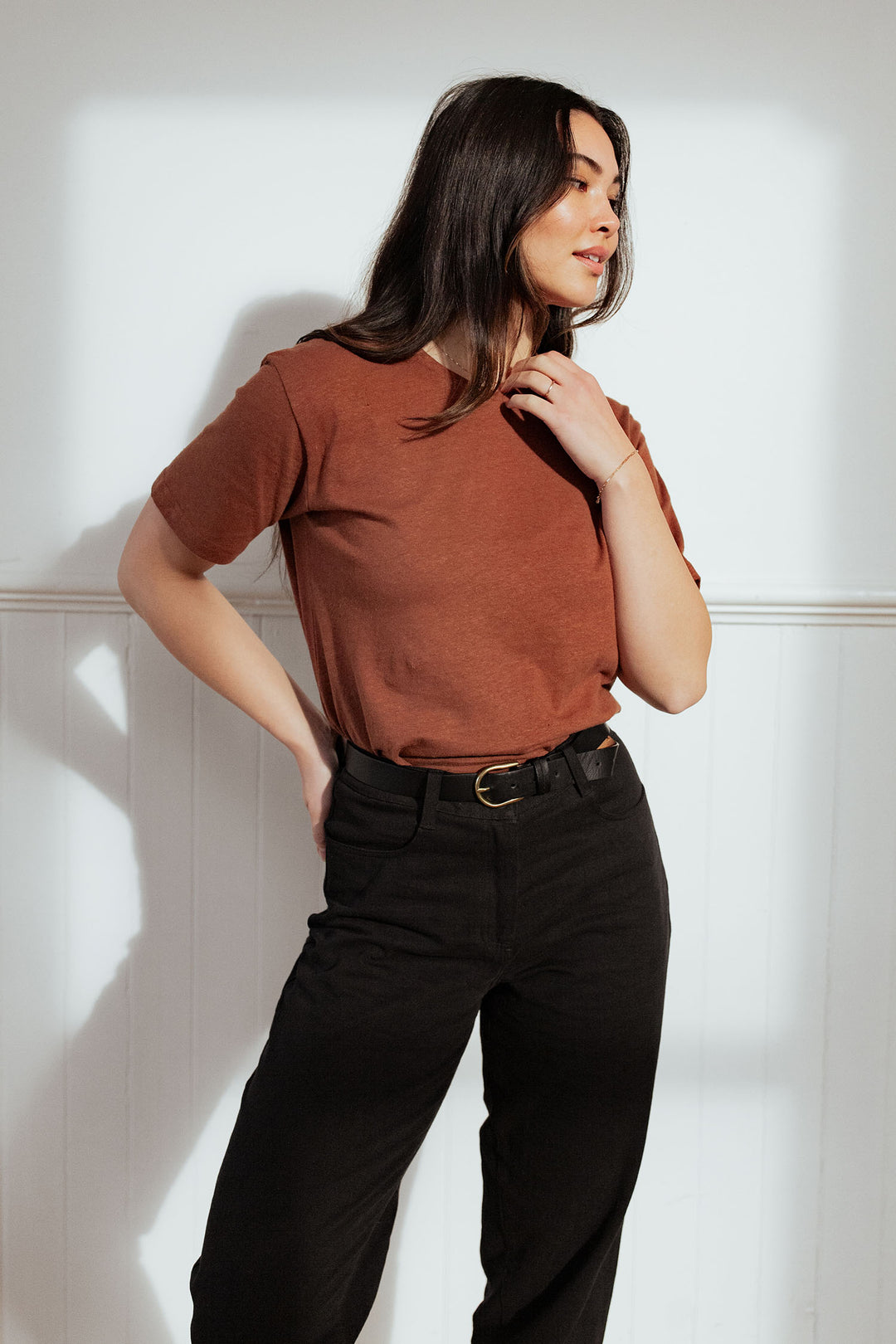 A woman with long dark hair models the Bridge & Burn Organic Hemp Boxy Tee in Burnt Umber with black pants, standing against a white wall and looking to the side, one hand on her neck and the other on her hip.