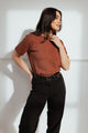 A woman with long dark hair models the Bridge & Burn Organic Hemp Boxy Tee in Burnt Umber with black pants, standing against a white wall and looking to the side, one hand on her neck and the other on her hip.