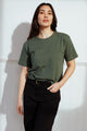 A person with long dark hair stands indoors against a white wall, posing confidently with one hand in their pocket and wearing the Bridge & Burn Organic Hemp Boxy Tee in Cypress and black pants.