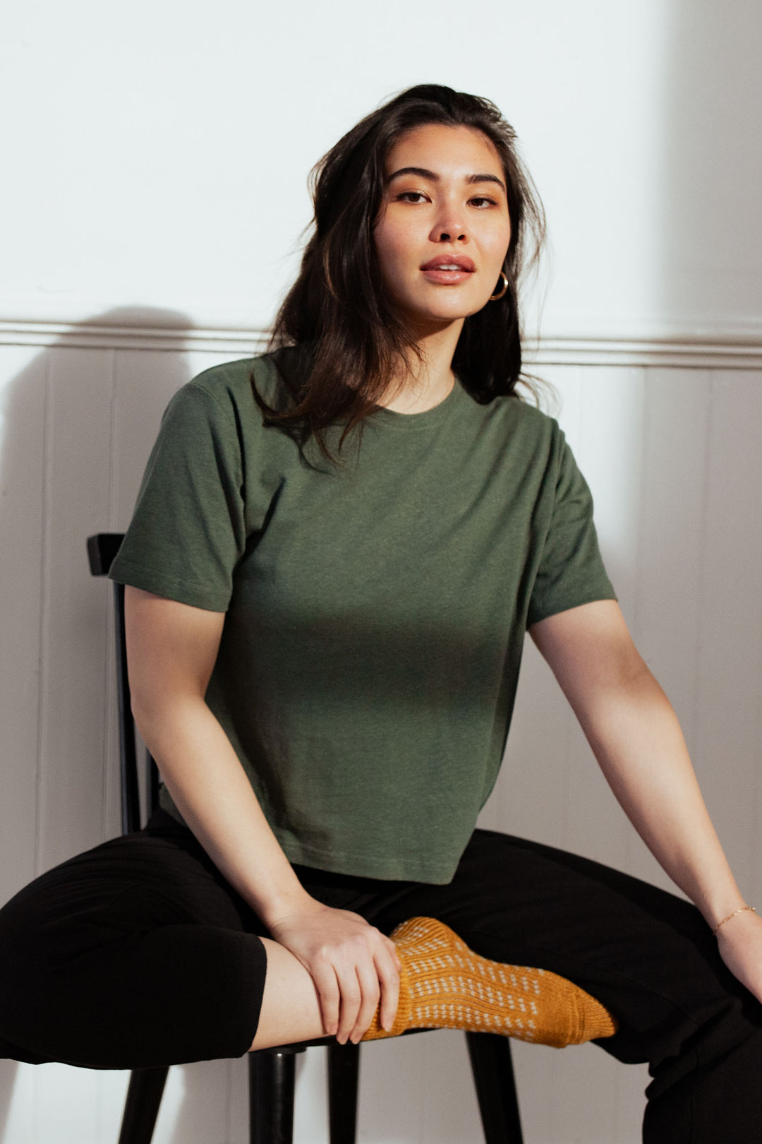 A woman with long dark hair sits on a chair wearing the Bridge & Burn Organic Hemp Boxy Tee in Cypress, black pants, and orange socks. She looks relaxed with one leg crossed and her hand resting on her knee, set against a white wall background.