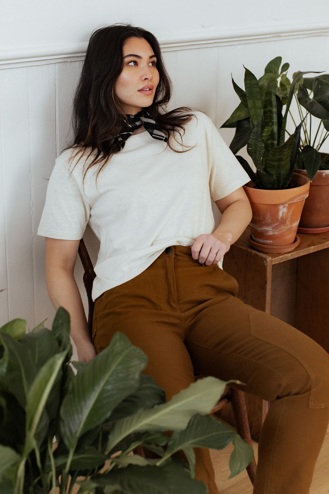 A woman with long dark hair sits on a chair near potted plants, wearing the Bridge & Burn Organic Hemp Boxy Tee in Ivory, brown pants, and a black-and-white scarf. She looks off to the side with a relaxed expression.