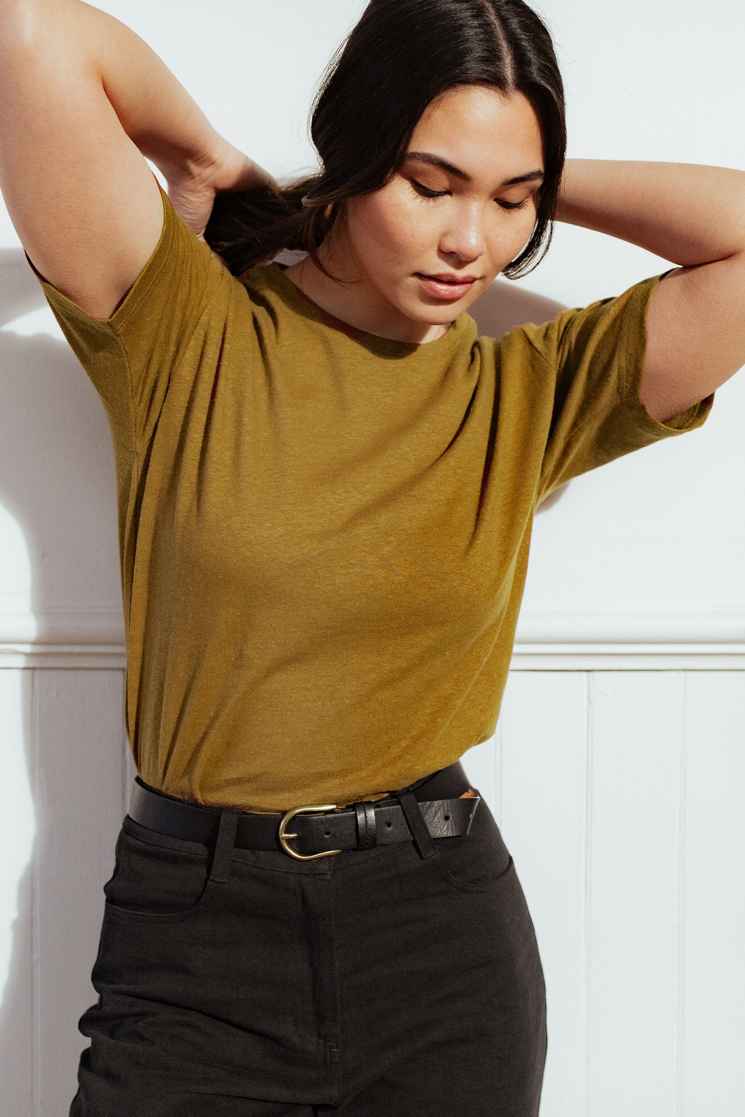 A person with long dark hair, wearing the Bridge & Burn Organic Hemp Boxy Tee in ochre and black pants, stands against a white wall, looking down and touching their hair with both hands.
