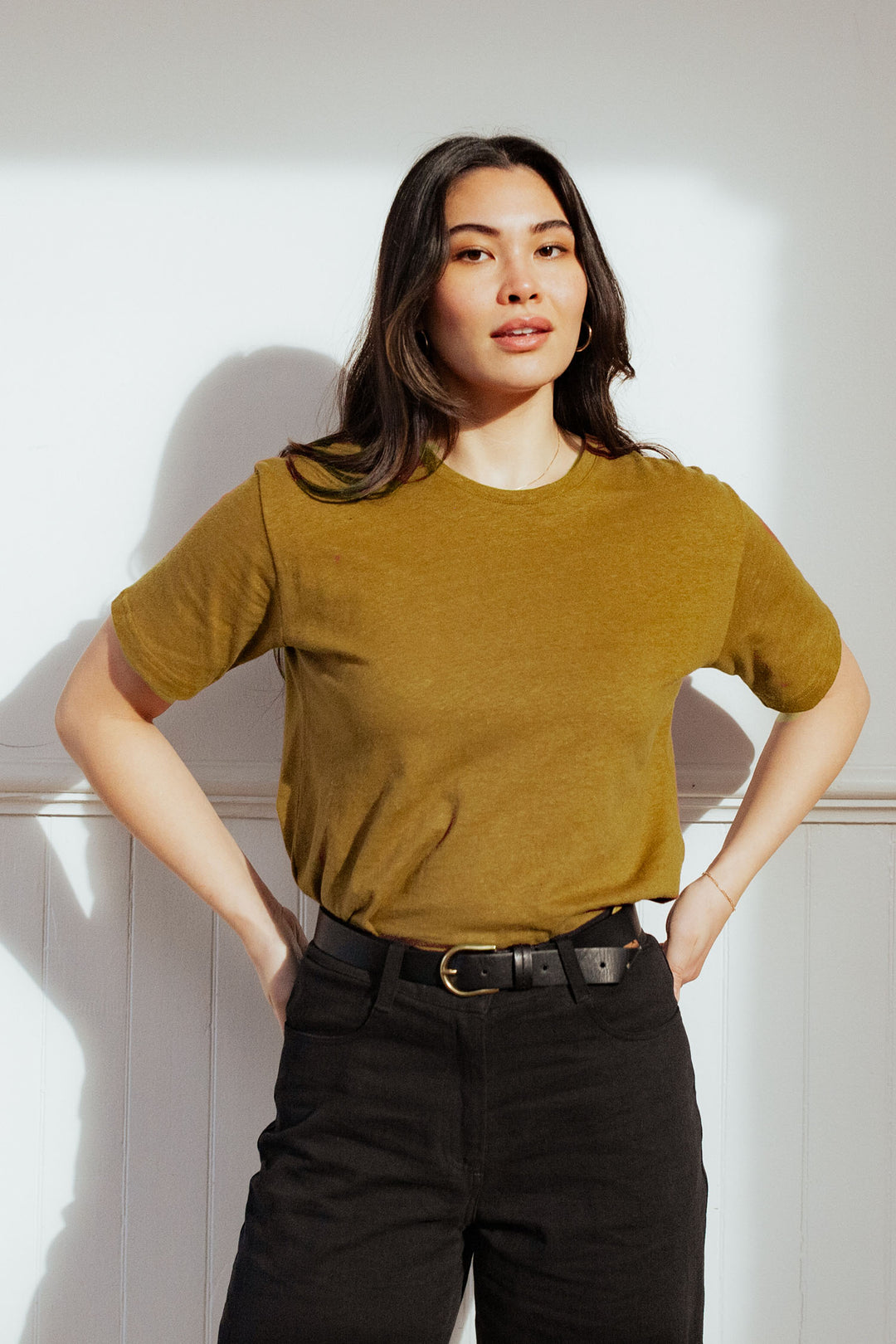 A woman with long dark hair stands confidently in front of a white wall, wearing the Bridge & Burn Organic Hemp Boxy Tee in Ochre, tucked into belted black pants, hands on her hips.