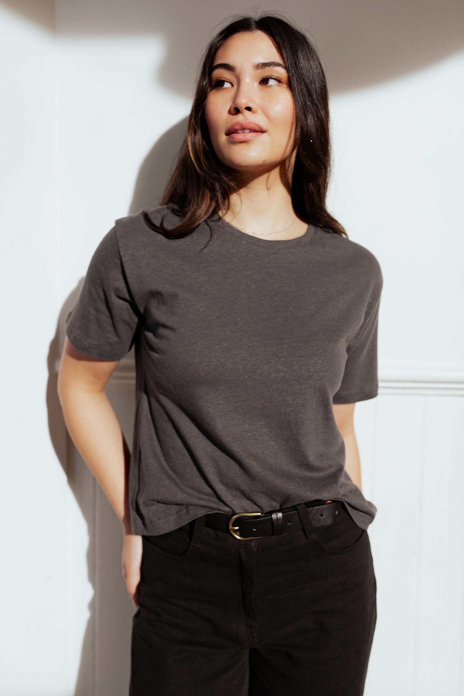 A woman with long dark hair stands against a white wall, wearing the Bridge & Burn Organic Hemp Boxy Tee in Slate Grey and black belted pants. She gazes upward, one hand in her pocket, radiating effortless style.