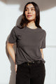 A woman with long dark hair stands against a white wall, wearing the Bridge & Burn Organic Hemp Boxy Tee in Slate Grey and black belted pants. She gazes upward, one hand in her pocket, radiating effortless style.