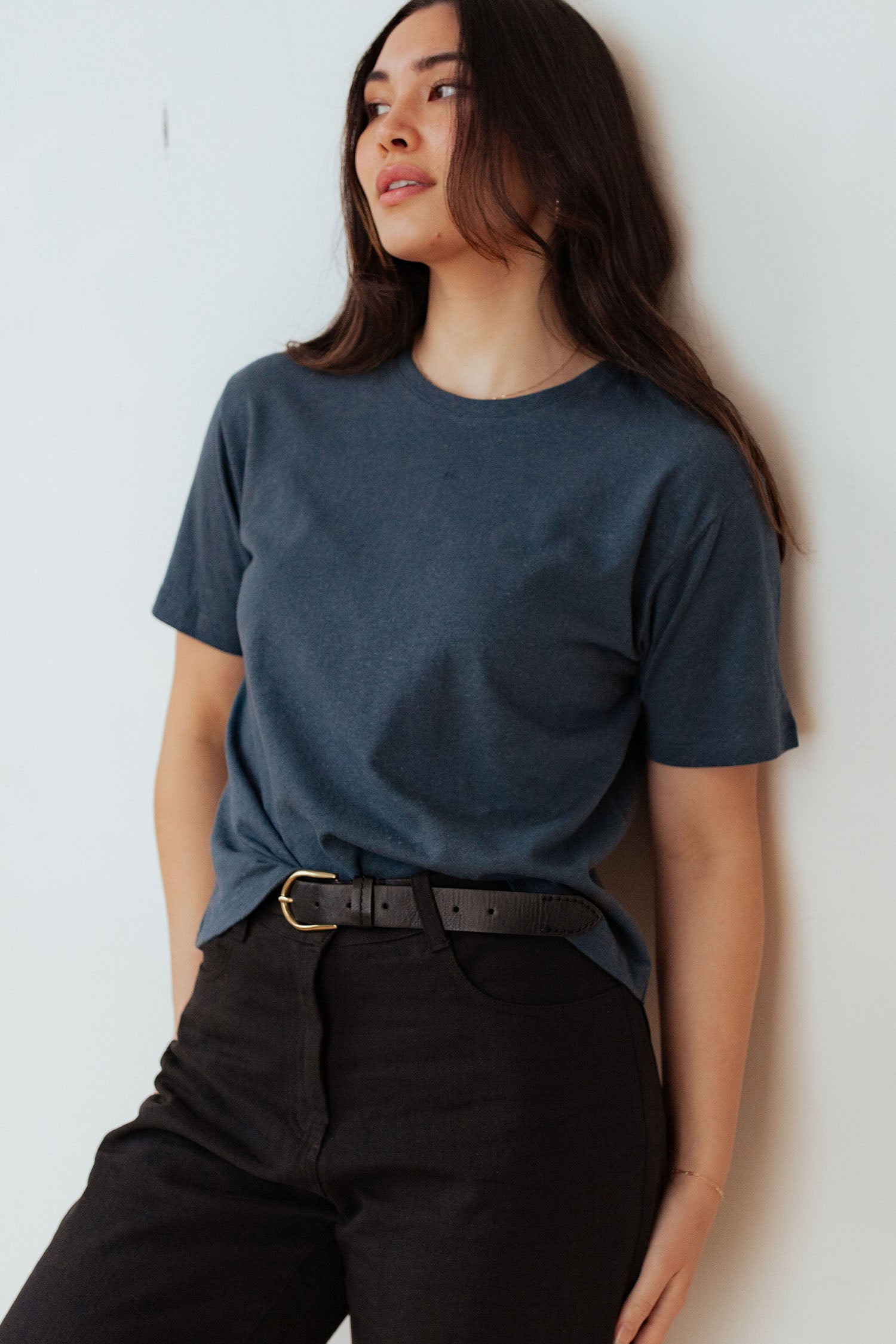A woman with long dark hair stands by a white wall, wearing the Bridge & Burn Organic Hemp Boxy Tee in Storm Blue, tucked into black pants with a black belt. She gazes to the side with a relaxed expression.