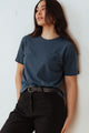 A woman with long dark hair stands by a white wall, wearing the Bridge & Burn Organic Hemp Boxy Tee in Storm Blue, tucked into black pants with a black belt. She gazes to the side with a relaxed expression.