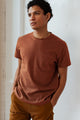 A person with short curly hair stands indoors against a light background, wearing Bridge & Burn's Organic Hemp Pocket Tee in Burnt Umber and brown pants, hands in their pockets and looking to the side.