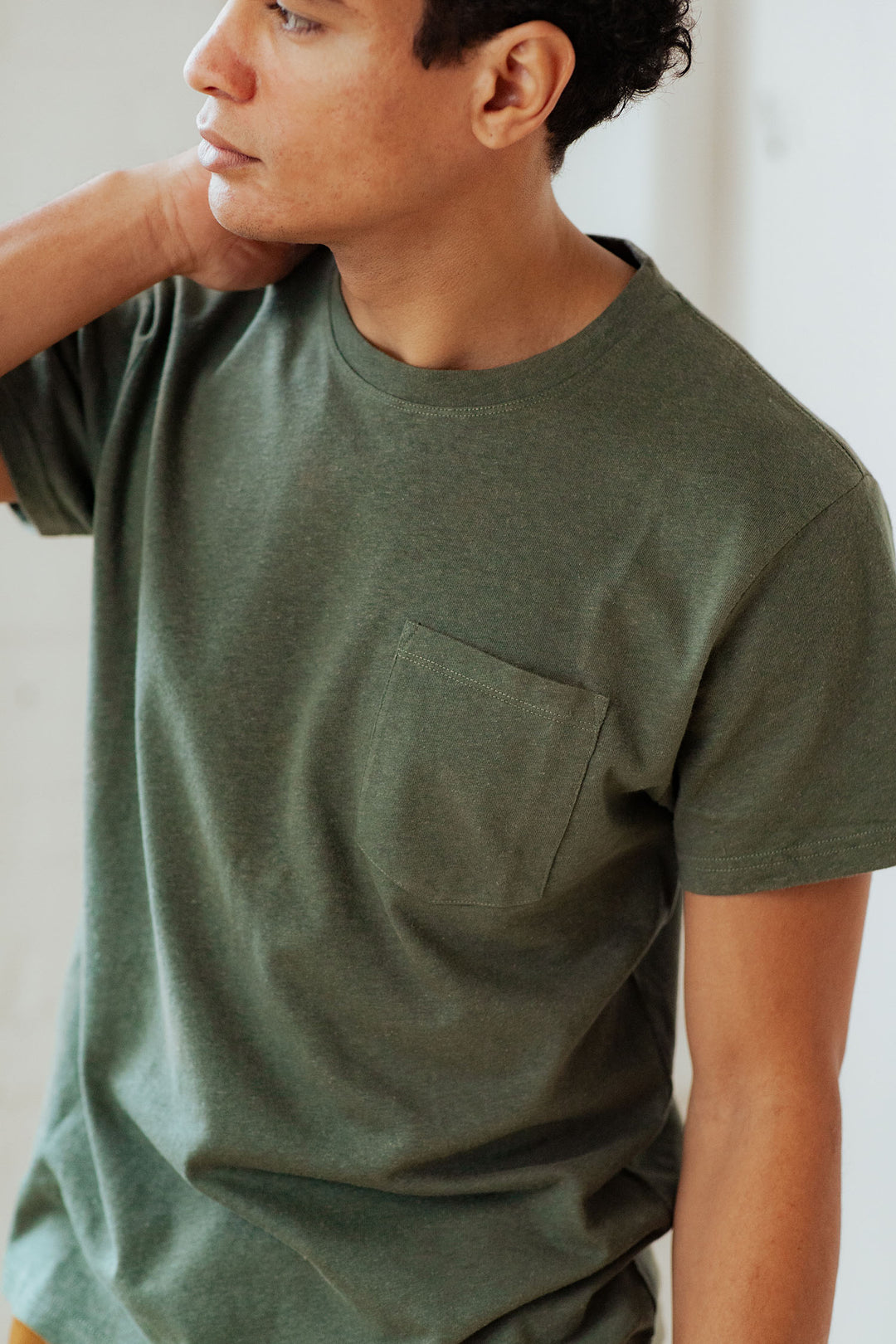 A person wears the Bridge & Burn Organic Hemp Pocket Tee in Cypress, featuring a chest pocket, and looks to the side with one hand on their neck. Only their upper body is shown.