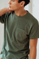 A person wears the Bridge & Burn Organic Hemp Pocket Tee in Cypress, featuring a chest pocket, and looks to the side with one hand on their neck. Only their upper body is shown.