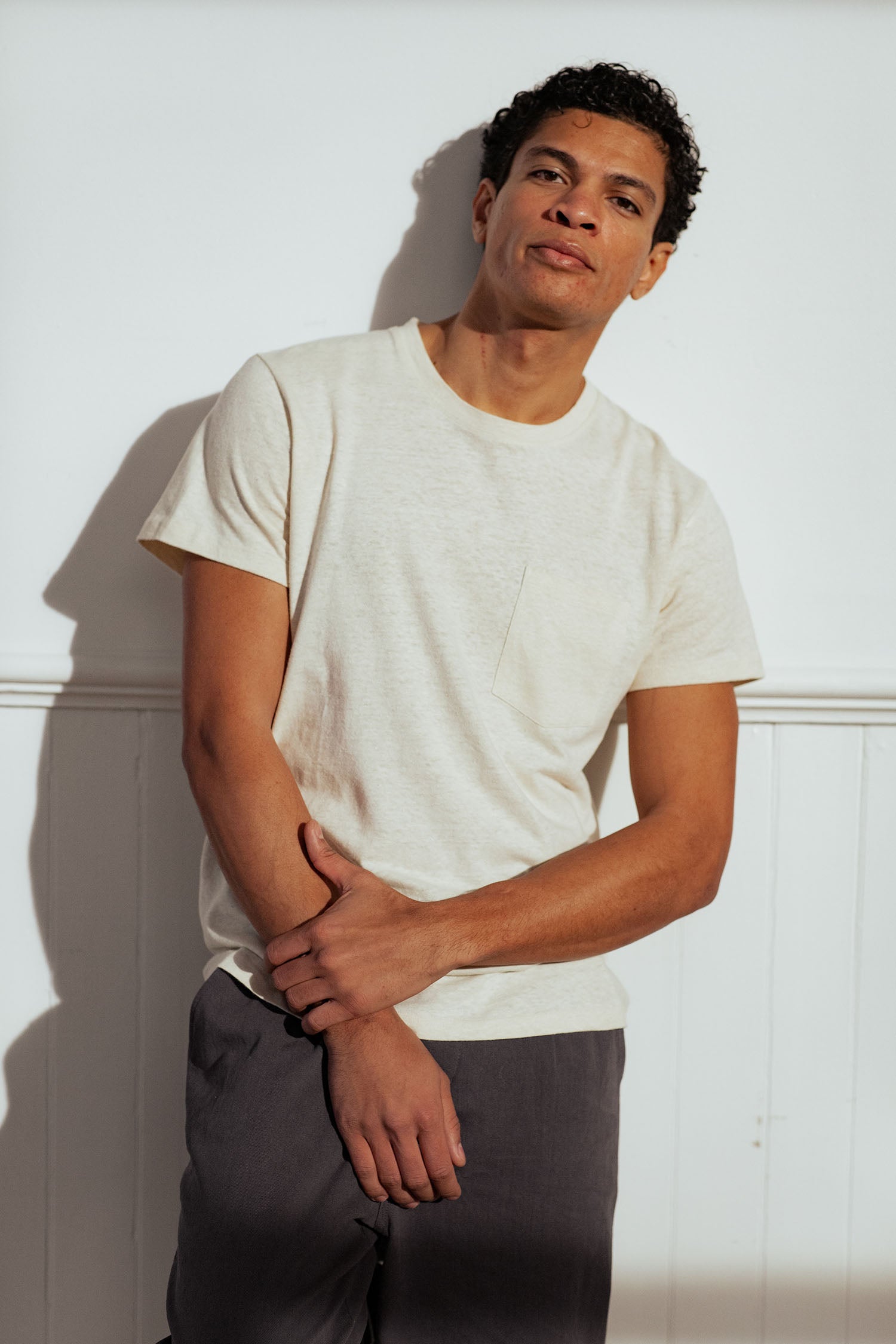 A man with short curly hair wears the Bridge & Burn Organic Hemp Pocket Tee in Ivory and dark pants, standing against a white wall and looking at the camera with a relaxed expression, one hand holding his other arm.
