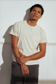 A man with short curly hair wears the Bridge & Burn Organic Hemp Pocket Tee in Ivory and dark pants, standing against a white wall and looking at the camera with a relaxed expression, one hand holding his other arm.