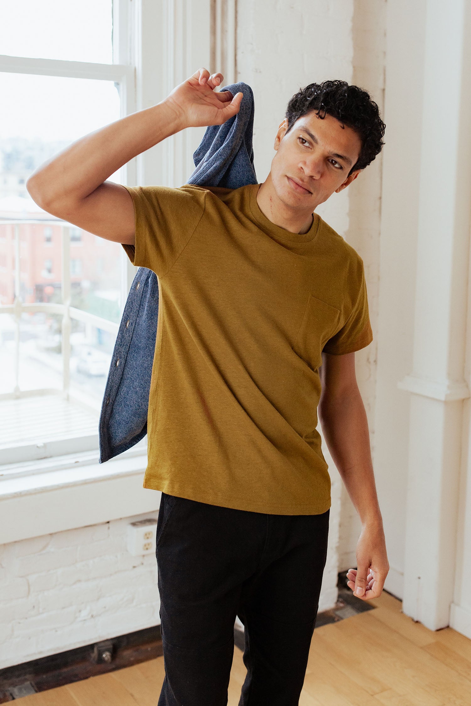 A man stands indoors by a window, wearing the Bridge & Burn Organic Hemp Pocket Tee in Ochre and black pants. He holds a blue jacket over his shoulder and gazes to the side with a relaxed expression.
