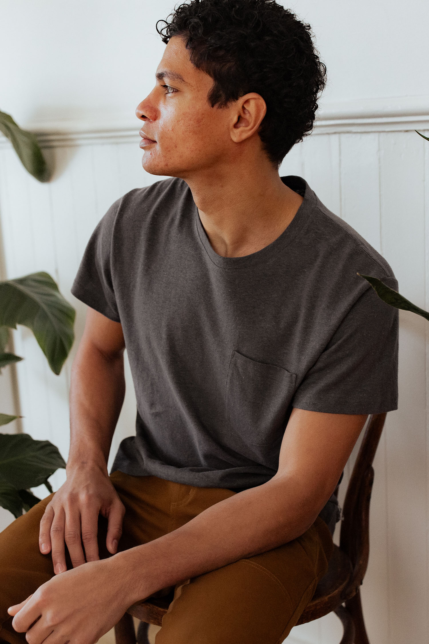 A man with curly hair sits on a wooden chair, wearing the Bridge & Burn Organic Hemp Pocket Tee in Slate Grey and mustard pants, looking to his left. Green plants and white paneled walls are in the background.