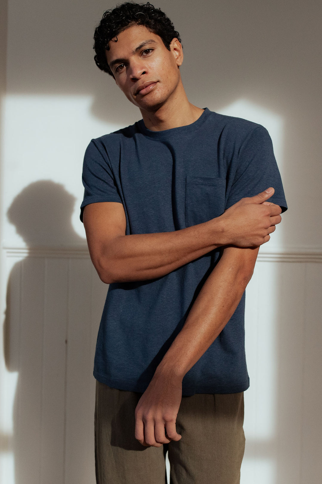 Standing in soft sunlight against a white wall, a person with short curly hair wears the Bridge & Burn Organic Hemp Pocket Tee in Storm Blue and brown pants, one arm crossed with a relaxed, thoughtful expression.