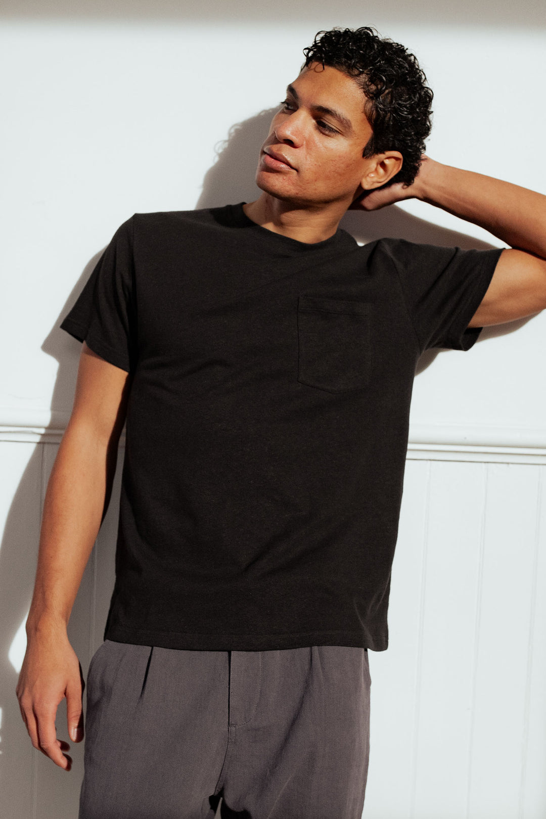 A man with curly hair stands against a white wall in Bridge & Burn’s Organic Hemp Pocket Tee in Washed Black and gray pants, posing with one hand behind his head to highlight the relaxed, sustainable style.