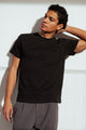 A man with curly hair stands against a white wall in Bridge & Burn’s Organic Hemp Pocket Tee in Washed Black and gray pants, posing with one hand behind his head to highlight the relaxed, sustainable style.