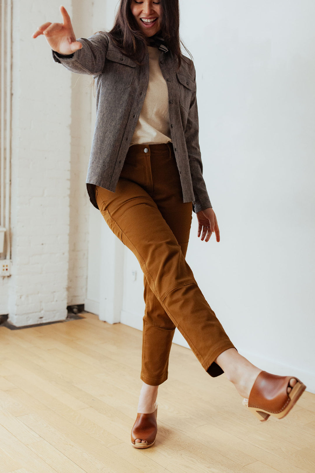 A woman smiles indoors, playfully lifting one leg while wearing Bridge & Burn's Bowden Utility Pant in Hickory Canvas with a cream top, gray jacket, and brown heeled sandals. The room features light wood floors and white walls.