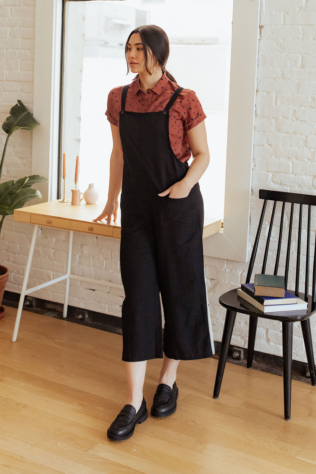 A woman stands indoors wearing the Edie Overall in Black by Bridge & Burn over a red short-sleeve shirt and black shoes. She is near a wooden desk adorned with candles and a vase, next to a black chair stacked with books.