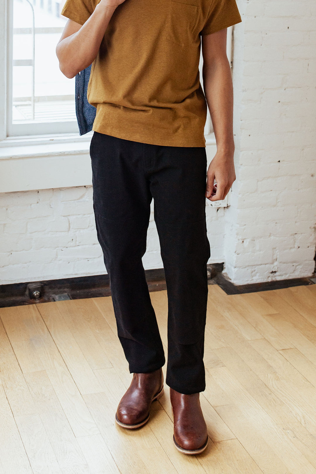 A person stands indoors on a wooden floor, wearing a mustard t-shirt, brown leather boots, and Bridge & Burn's Wallace Utility Pant in Black Canvas. Their head is not visible in the image.