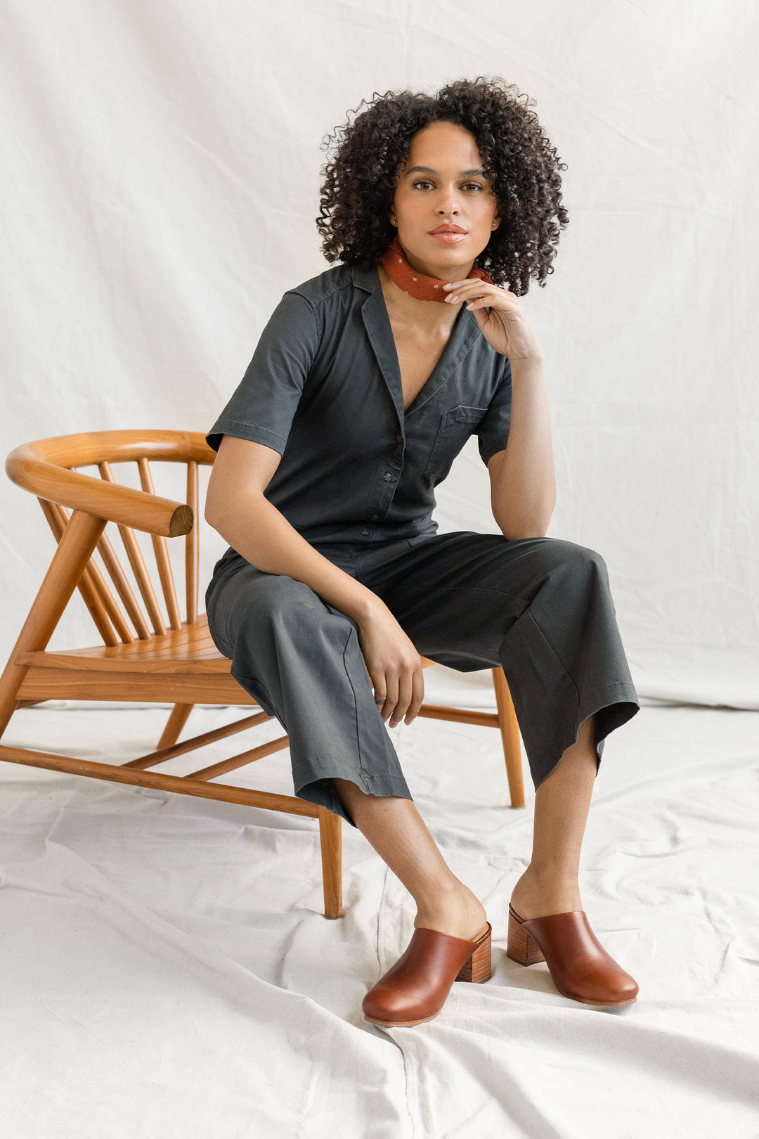 A person with curly hair sits on a wooden chair, wearing the Bridge & Burn Lou Utility Jumpsuit in Slate, paired with brown heeled clogs and a red bandana, set against a light, fabric-draped background.