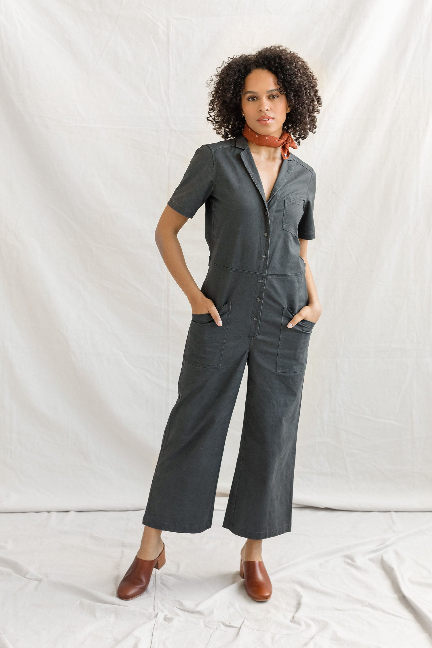 A woman with curly hair stands against a white backdrop, wearing the Bridge & Burn Lou Utility Jumpsuit in Slate, styled with brown heeled shoes and a red scarf, her hands resting in her pockets.