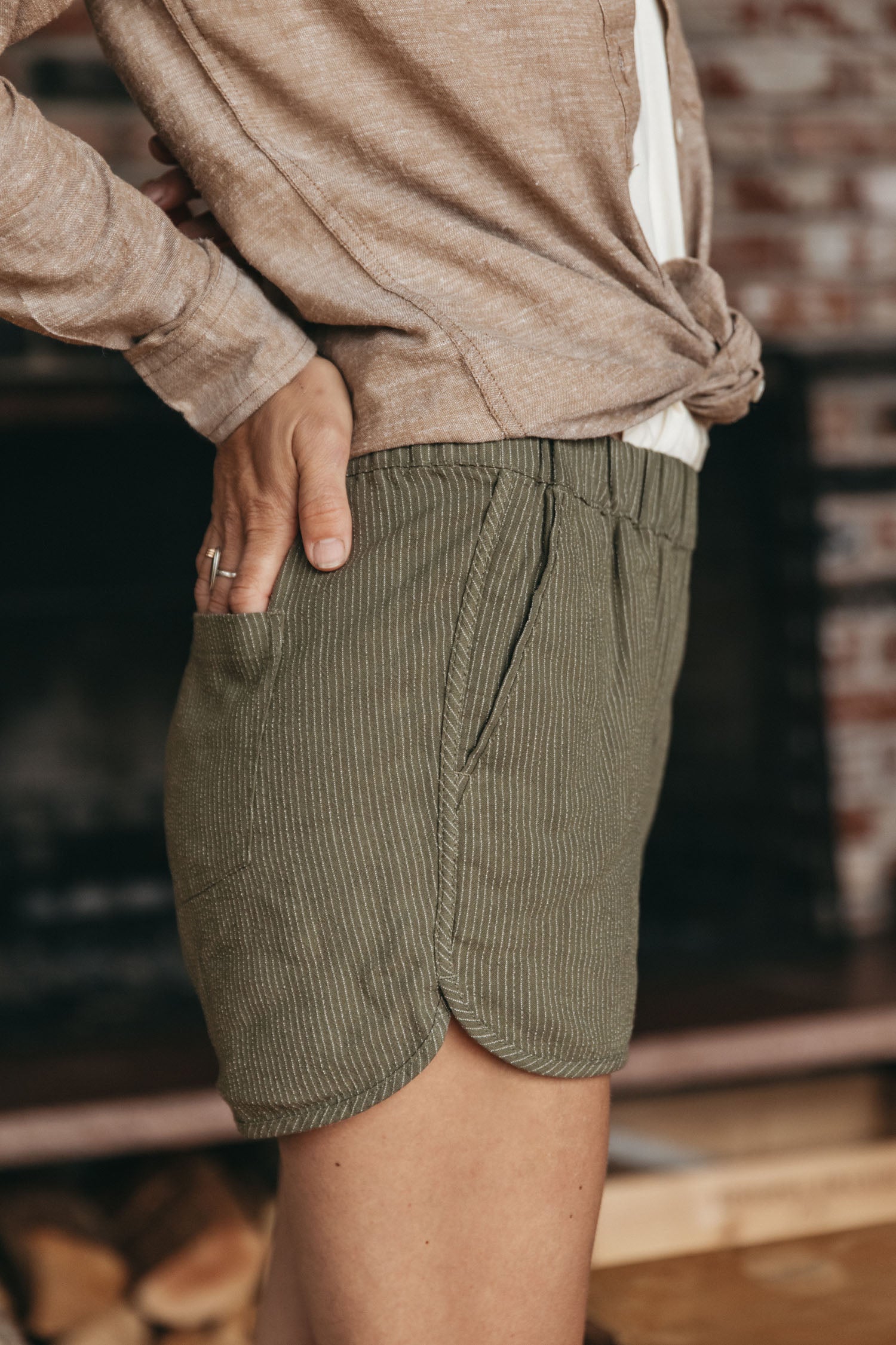 Wearing the Bridge & Burn Luca Track Short in Olive Pinstripe, paired with an oatmeal sweater and white shirt, a person stands indoors with one hand in a pocket; only their side, torso, and legs are visible.