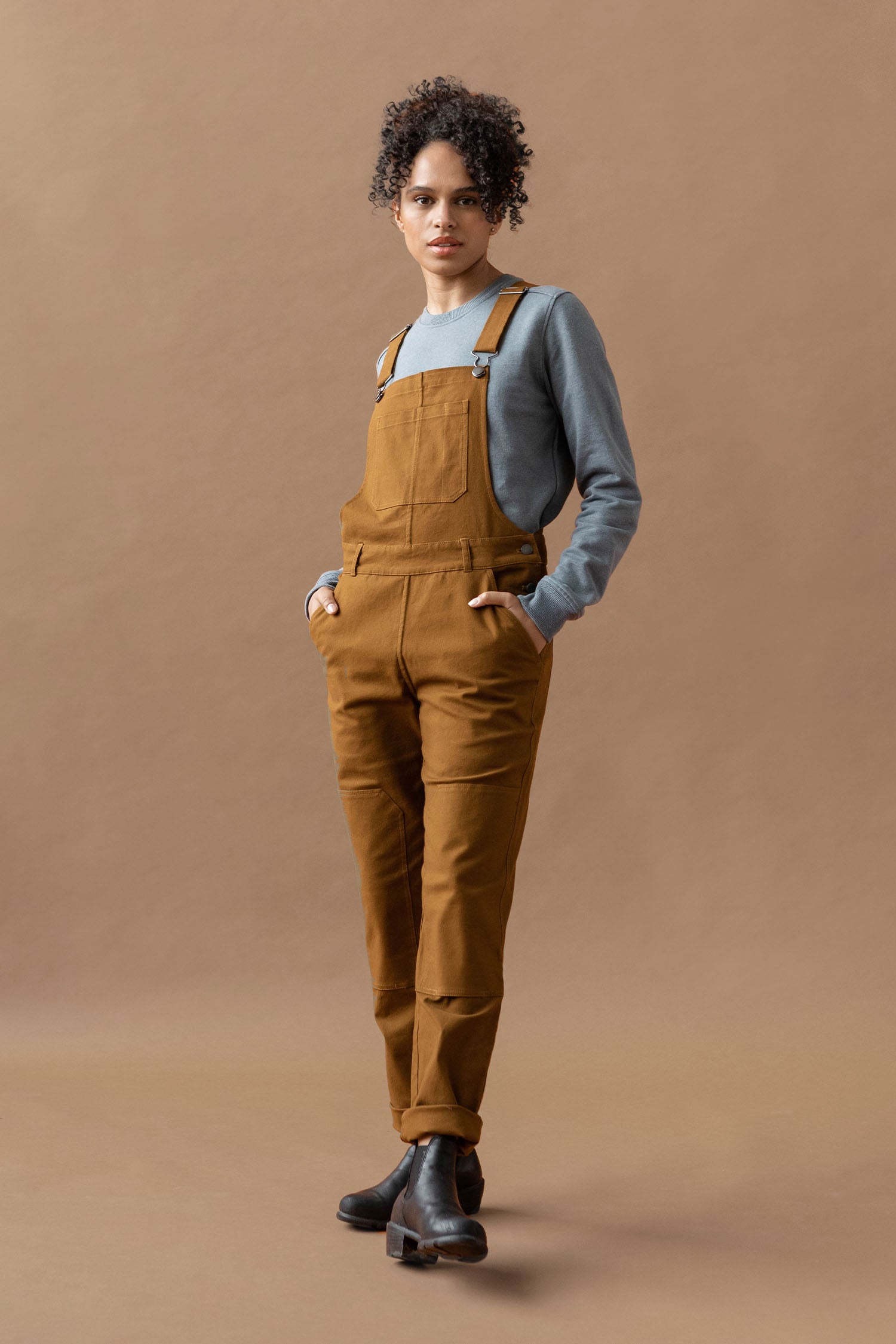 A person with short curly hair stands against a brown background wearing the Bridge & Burn Tobin Utility Overall in brown canvas and black boots, hands in pockets and a neutral expression.
