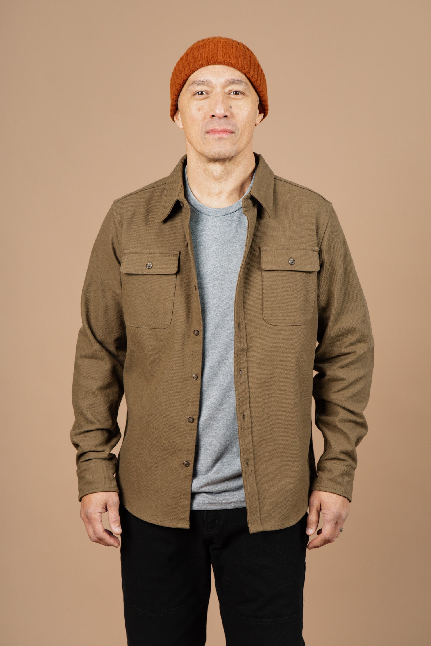 A person wears the Bridge & Burn Bedford Shirt in olive twill over a gray t-shirt, black pants, and an orange knit beanie, standing against a plain beige background and looking directly at the camera.