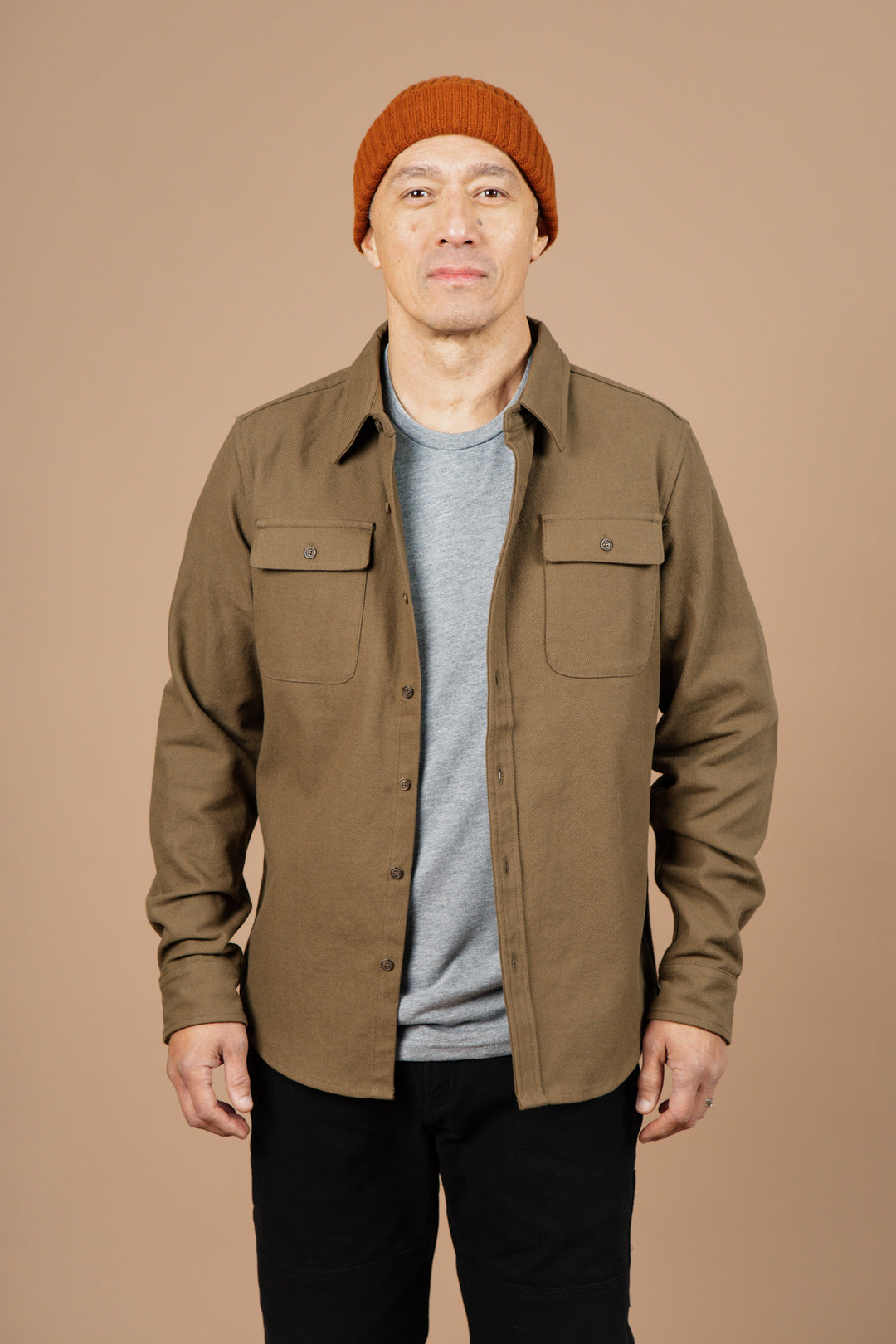 A person wears the Bridge & Burn Bedford Shirt in olive twill over a gray t-shirt, black pants, and an orange knit beanie, standing against a plain beige background and looking directly at the camera.
