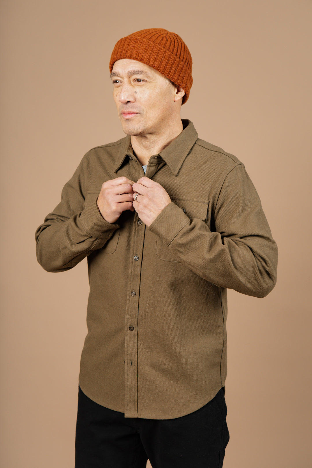 A man wearing the Bridge & Burn Bedford Shirt in Olive Twill and an orange knit beanie stands against a plain beige background, looking to the side while buttoning his shirt.