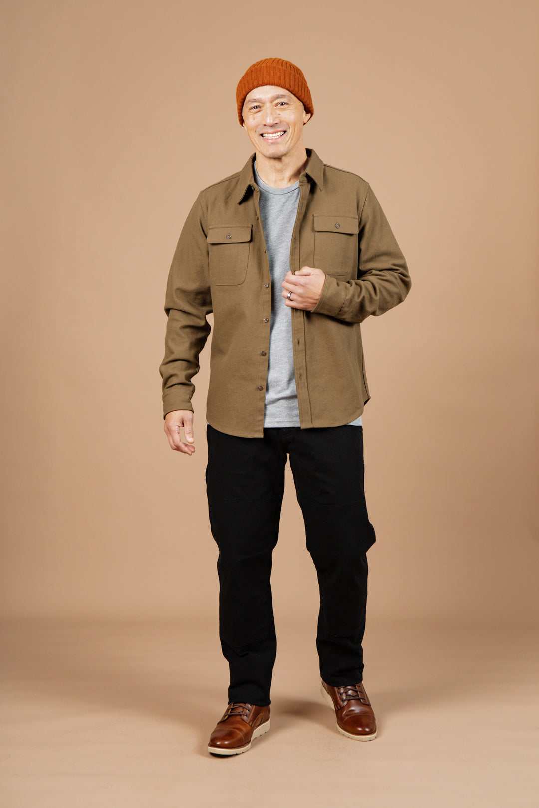A man in a brown beanie, gray t-shirt, black pants, brown shoes, and a Bridge & Burn Bedford Shirt in Olive Twill smiles against a plain beige background.