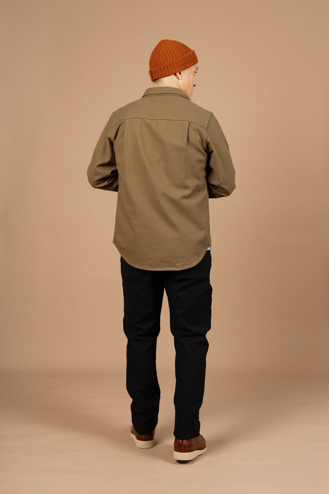 A person stands with their back to the camera, wearing the Bridge & Burn Bedford Shirt in Olive Twill, paired with black pants and brown shoes, set against a plain beige background.