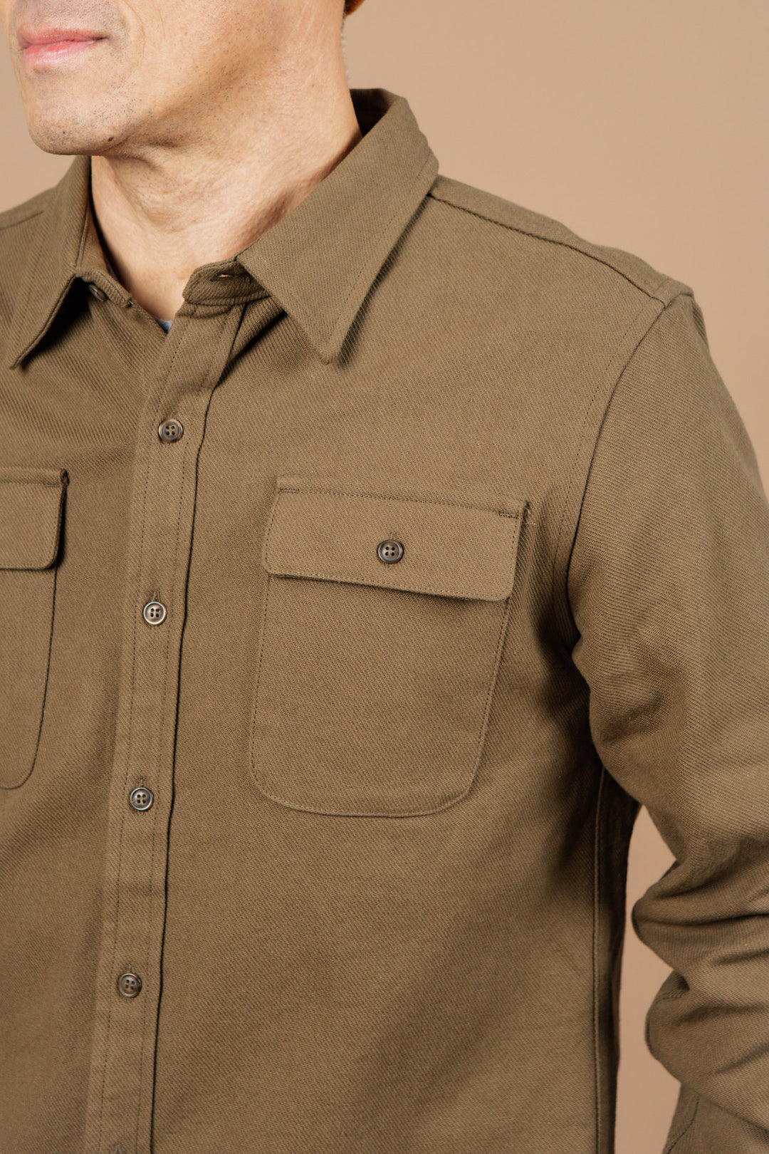 A man wearing the Bridge & Burn Bedford Shirt in Olive Twill, featuring two front pockets, stands against a beige background with only his lower face and upper torso visible.