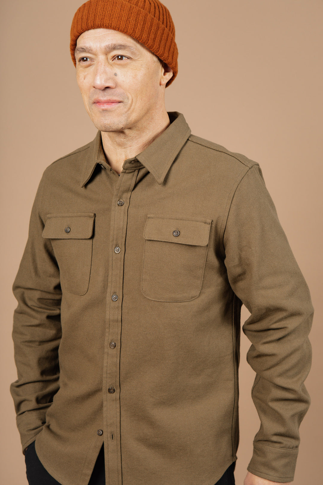 A man wearing the Bridge & Burn Bedford Shirt in Olive Twill with two chest pockets and an orange knit beanie stands against a beige background, looking to the side with a gentle smile.
