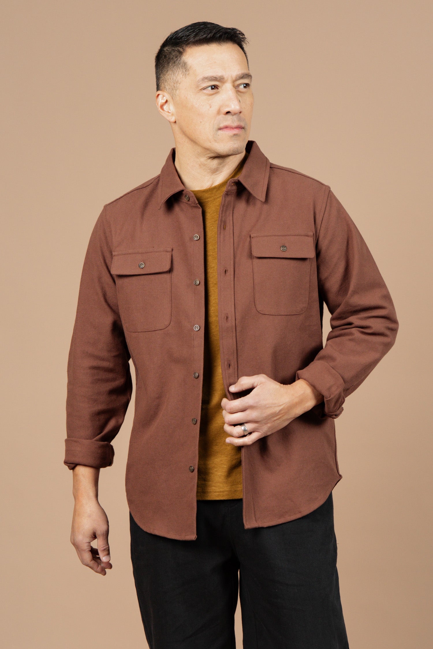A man with short dark hair wears the Bridge & Burn Bedford Shirt in Penny Twill over a tan tee and black pants, standing against a plain beige background and gazing slightly to his left.