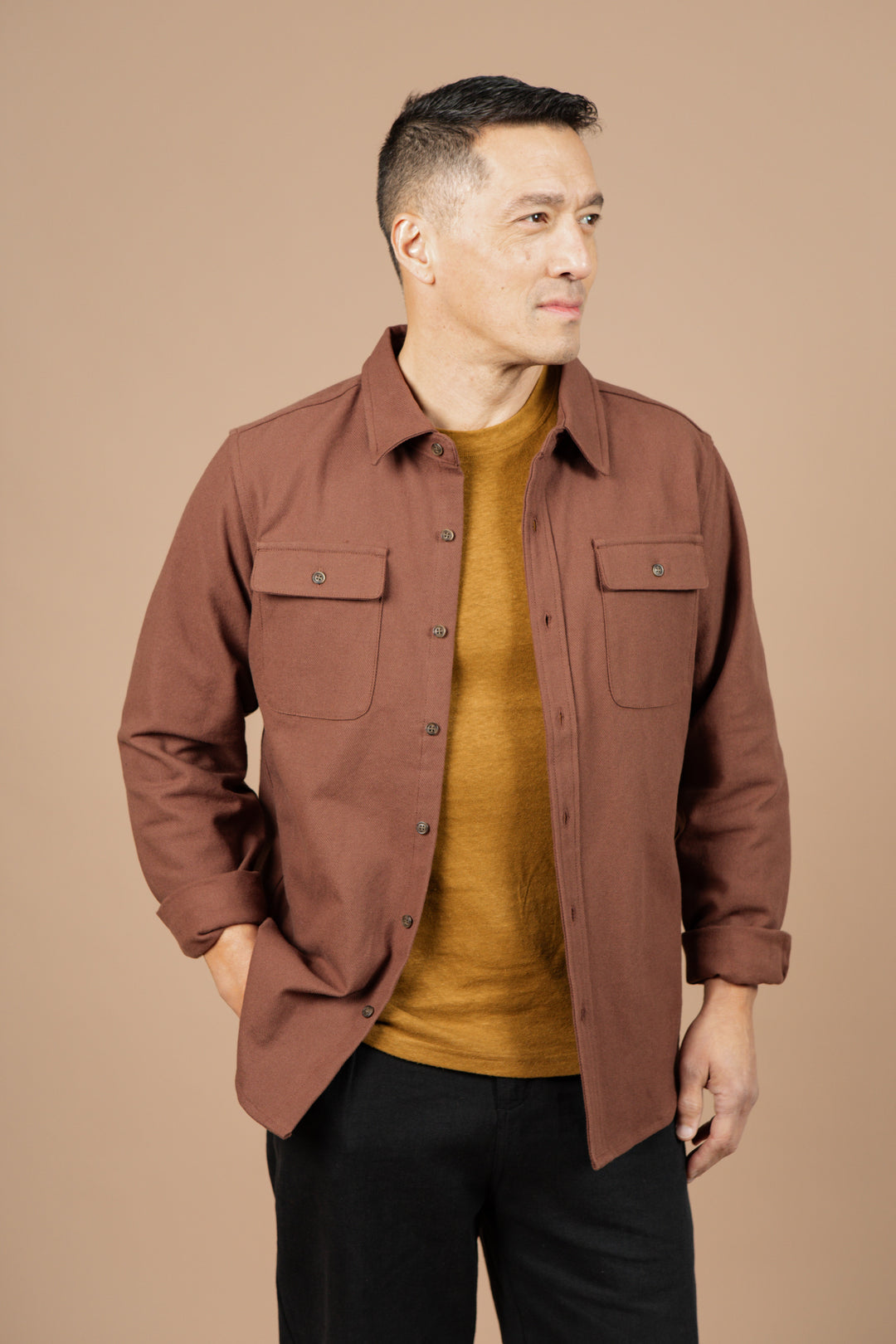 A man with short dark hair wears the Bridge & Burn Bedford Shirt in Penny Twill over a mustard yellow tee and black pants, standing against a beige background and looking to the side with one hand in his pocket.