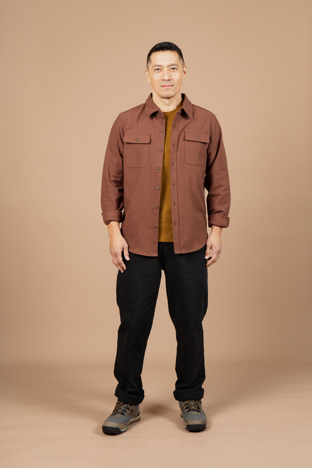 A man faces forward against a beige background, wearing Bridge & Burn’s Bedford Shirt in Penny Twill over a mustard shirt, paired with black pants and gray hiking shoes.