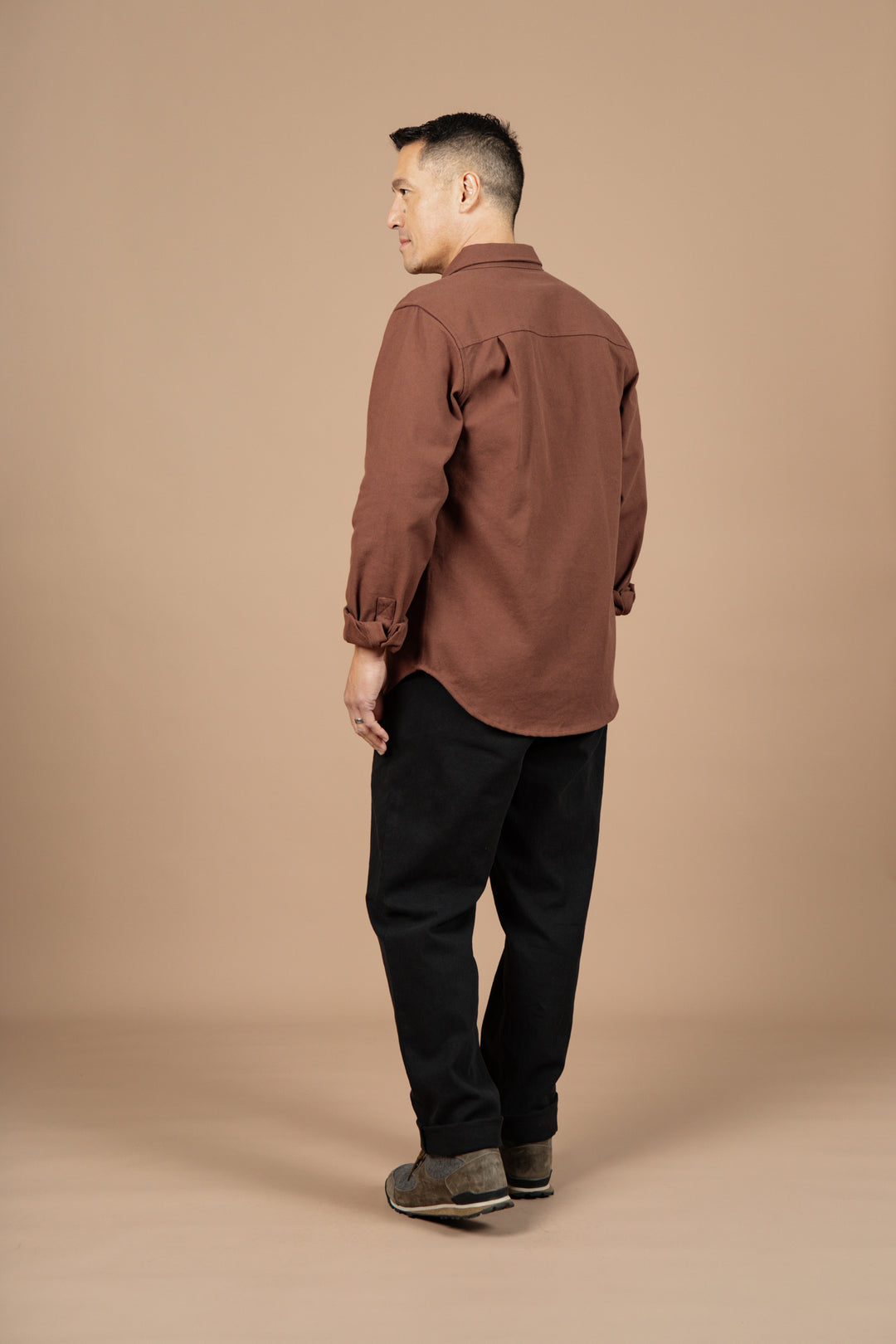 A man stands at an angle, wearing the Bridge & Burn Bedford Shirt in Penny Twill, paired with black pants and brown shoes against a plain beige background.