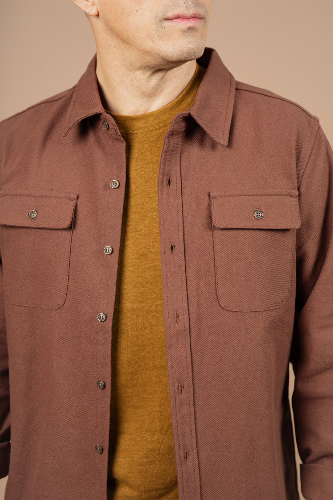 A man wears the Bridge & Burn Bedford Shirt in Penny Twill, a brown cotton twill shirt with chest pockets and standard fit, layered over a mustard yellow crew neck. He stands against a beige background, his face partially out of frame.