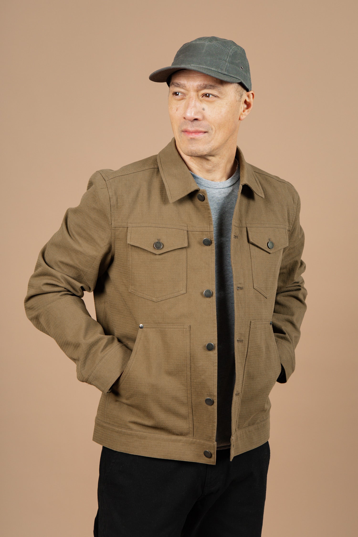 A man wearing the Bridge & Burn Burl Trucker Jacket in Warm Olive stands with his hands in his jacket pockets against a plain beige background.