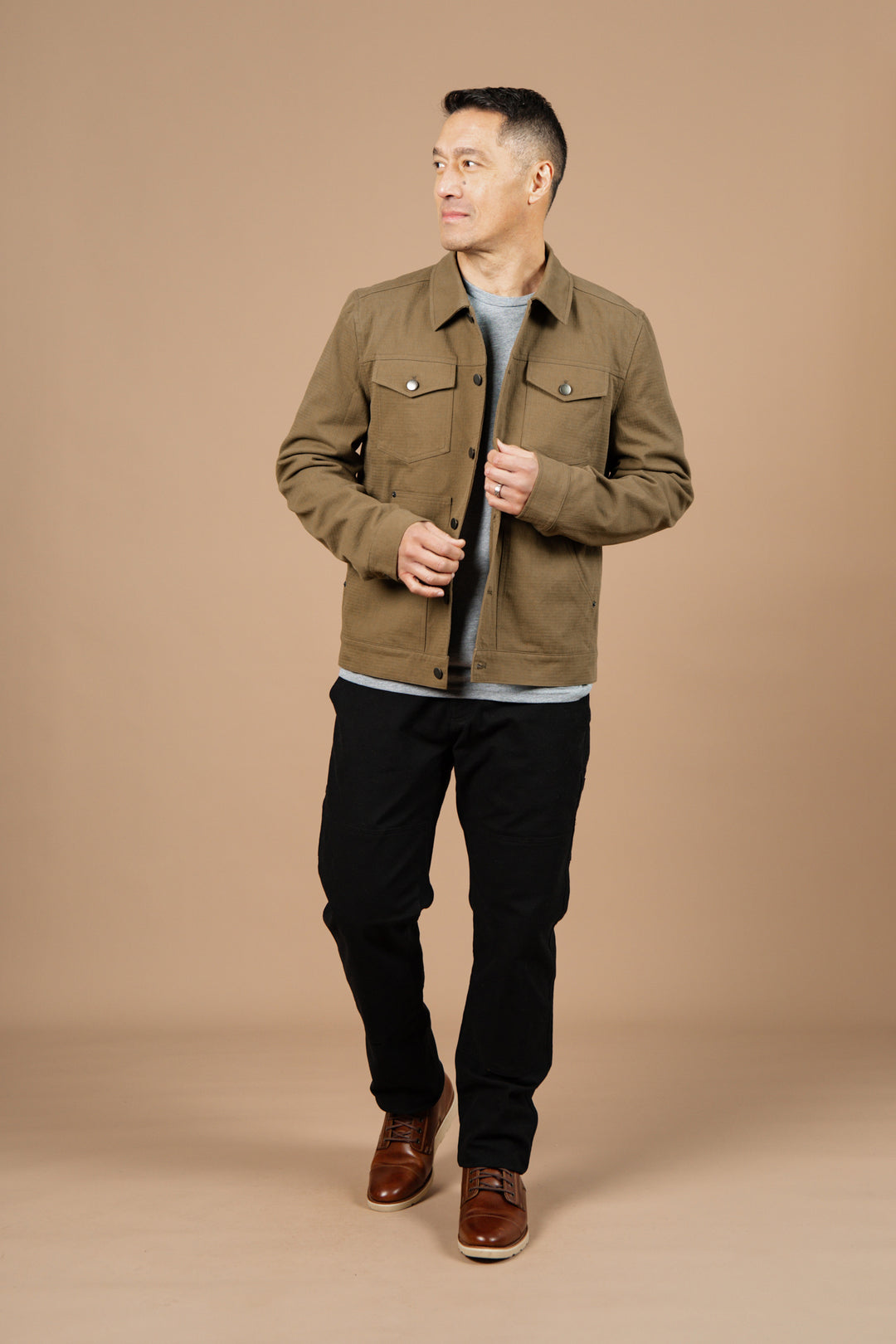 A man wears the Bridge & Burn Burl Trucker Jacket in Warm Olive over a gray shirt with black pants and brown shoes, standing against a plain beige background and looking slightly to the side with a neutral expression.