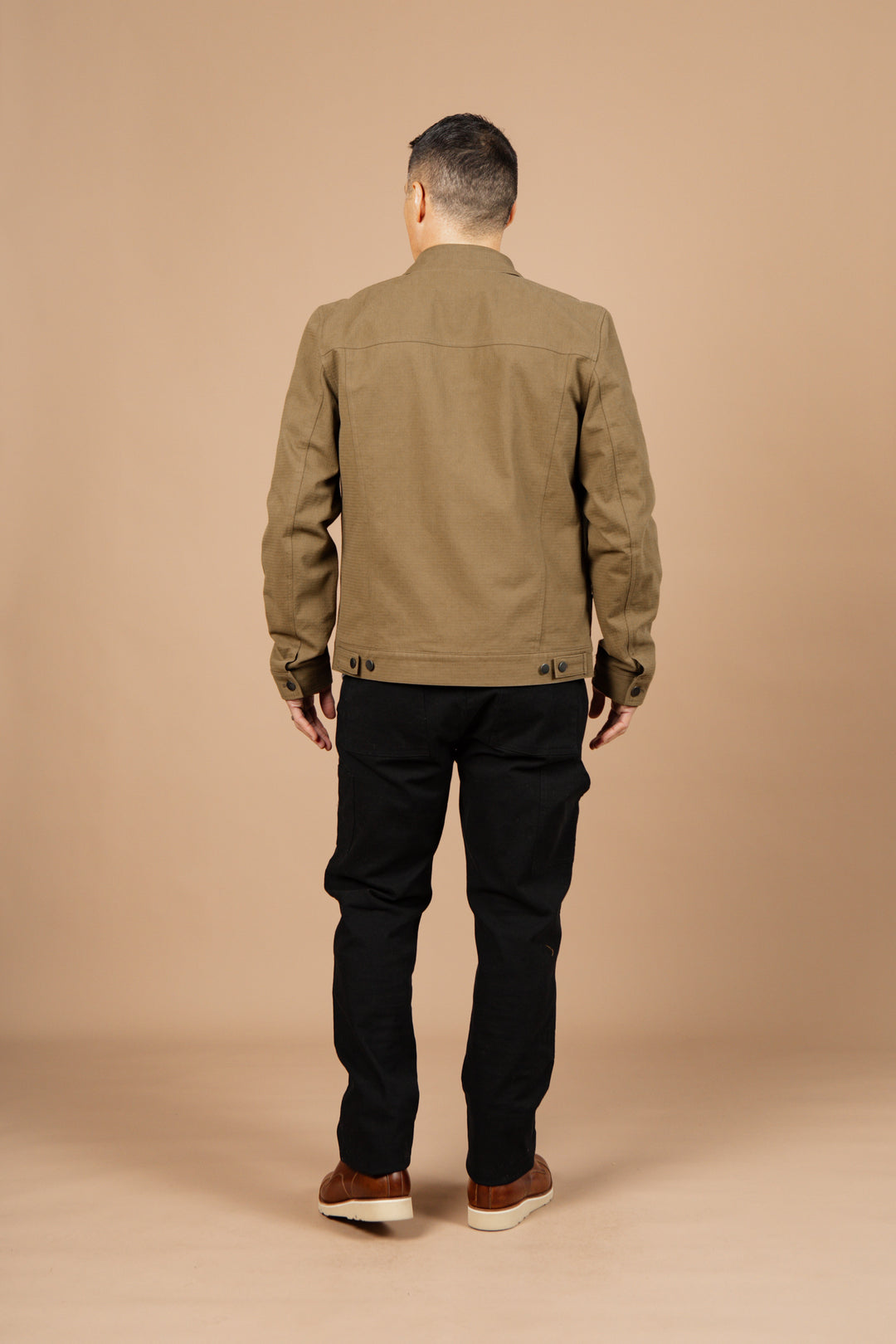 A man faces away from the camera against a beige background, wearing Bridge & Burn's Burl Trucker Jacket in Warm Olive, paired with black pants and brown shoes.