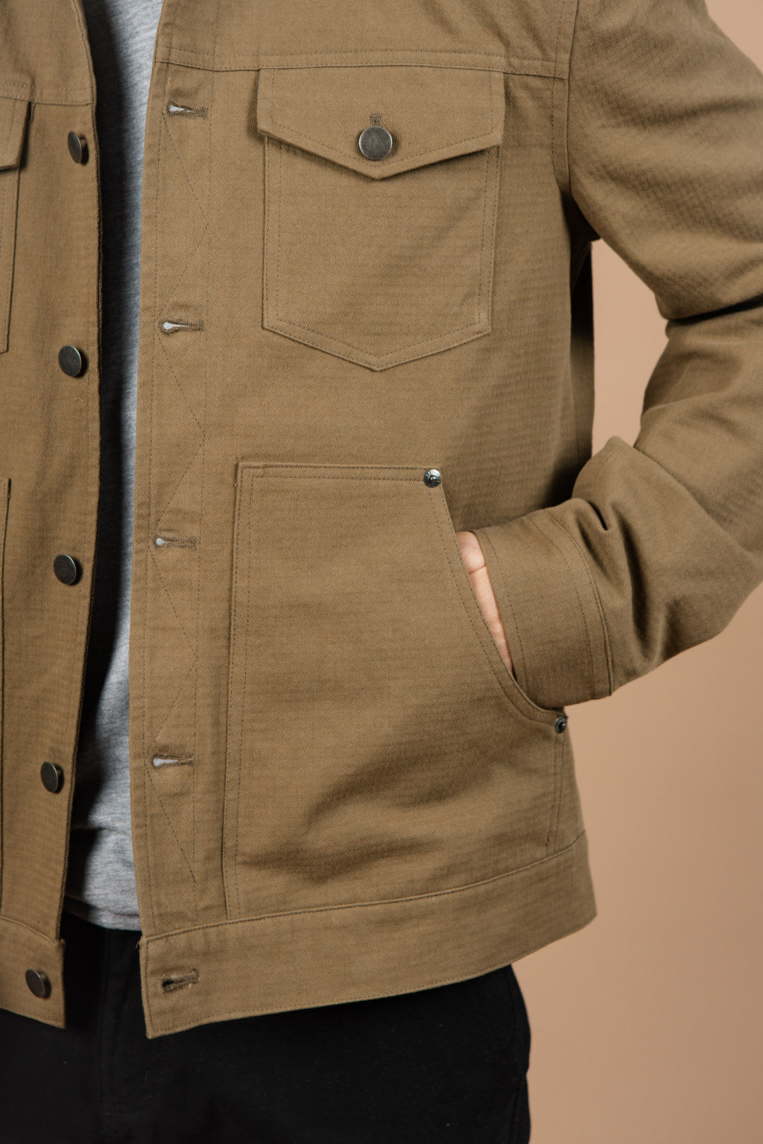 A person wears the Bridge & Burn Burl Trucker Jacket in Warm Olive, featuring a flap chest pocket and side hand pocket, standing against a beige background with their right hand in the side pocket. Only the torso and part of the arms are shown.