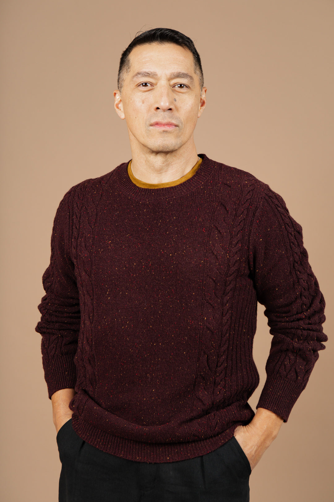 A man with short dark hair stands facing the camera against a plain beige background, wearing the Bridge & Burn Hugo Sweater in Burgundy Donegal with dark pants and his hands in his pockets.