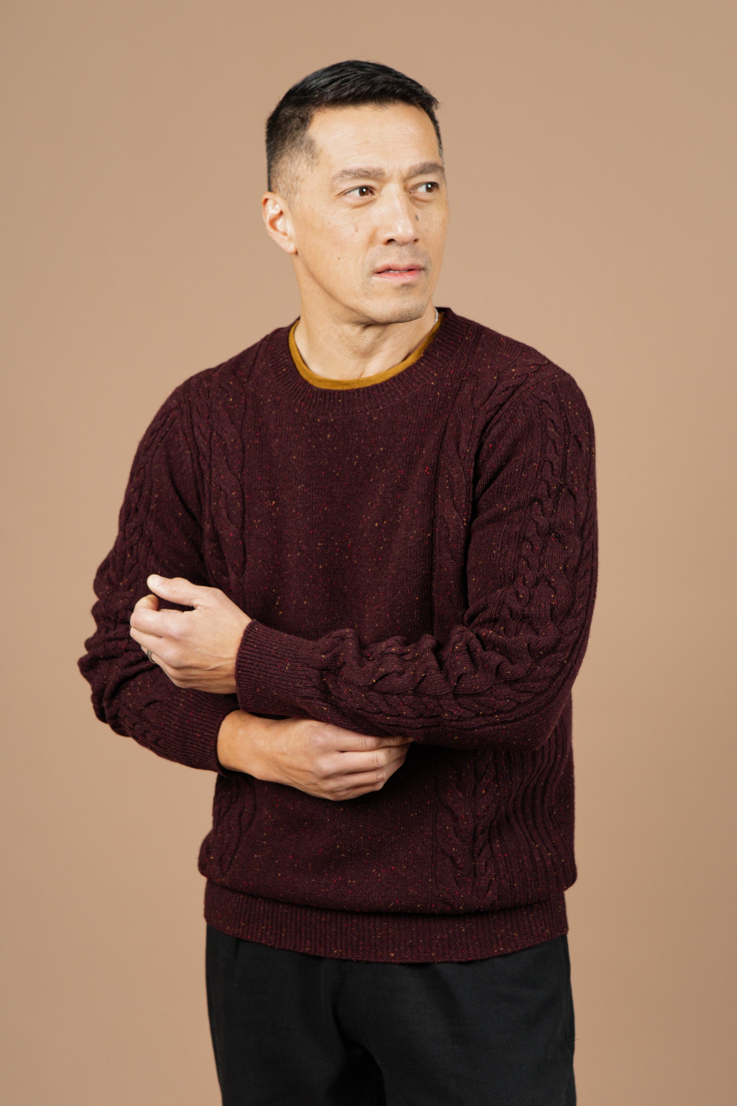 A man wears the Bridge & Burn Hugo Sweater in Burgundy Donegal and black pants, standing against a beige background while looking right and adjusting his sleeve.