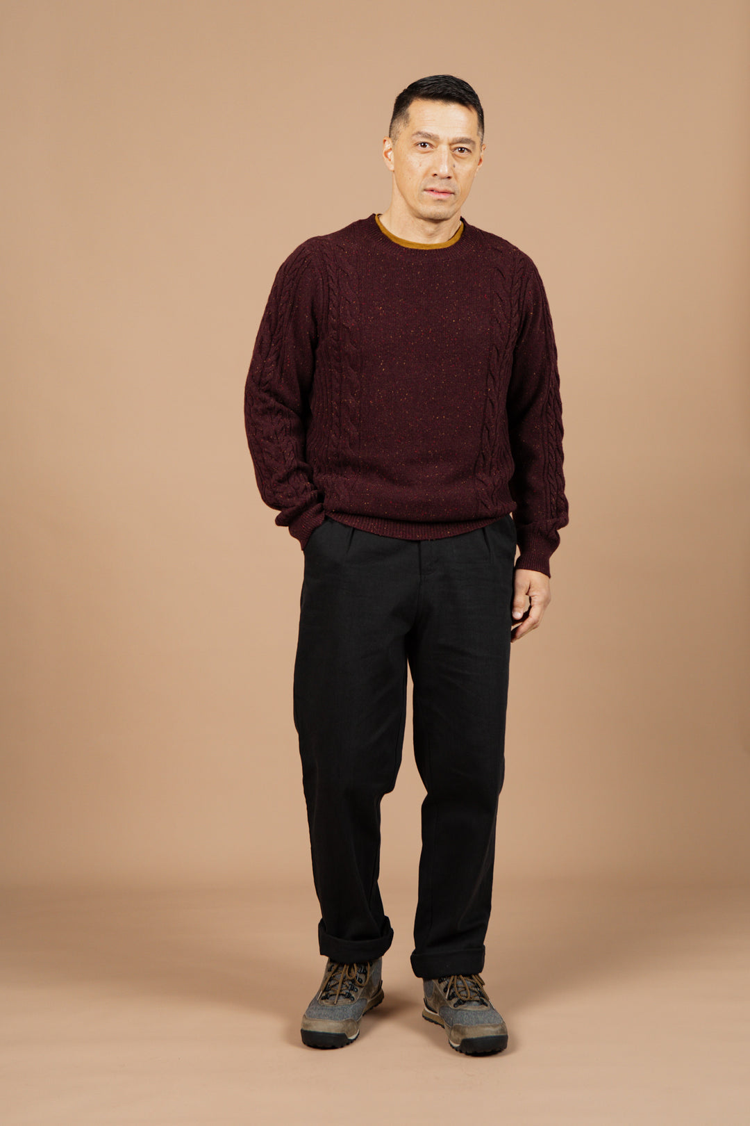 A man stands against a beige background wearing the Bridge & Burn Hugo Sweater in Burgundy Donegal, paired with black pants and gray shoes. He has short dark hair, a neutral expression, and one hand slightly in his pocket.