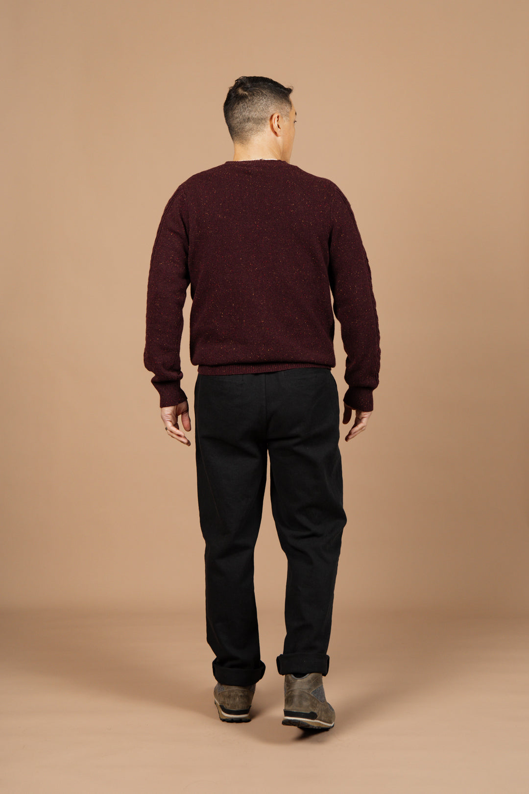 Facing away from the camera against a beige background, a person with short hair wears the Bridge & Burn Hugo Sweater in Burgundy Donegal, paired with black pants and brown shoes.
